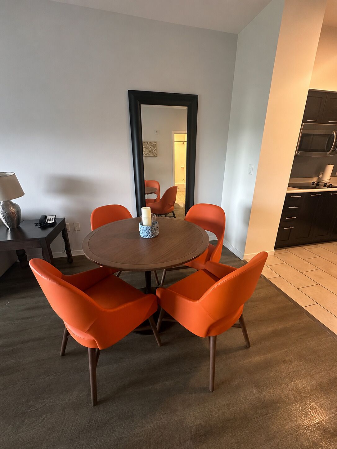 Un espace repas moderne avec une table ronde et des chaises orange, un miroir au mur et une cuisine dotée de meubles sombres.
