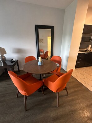 Un espace repas moderne avec une table ronde et des chaises orange, un miroir au mur et une cuisine dotée de meubles sombres.