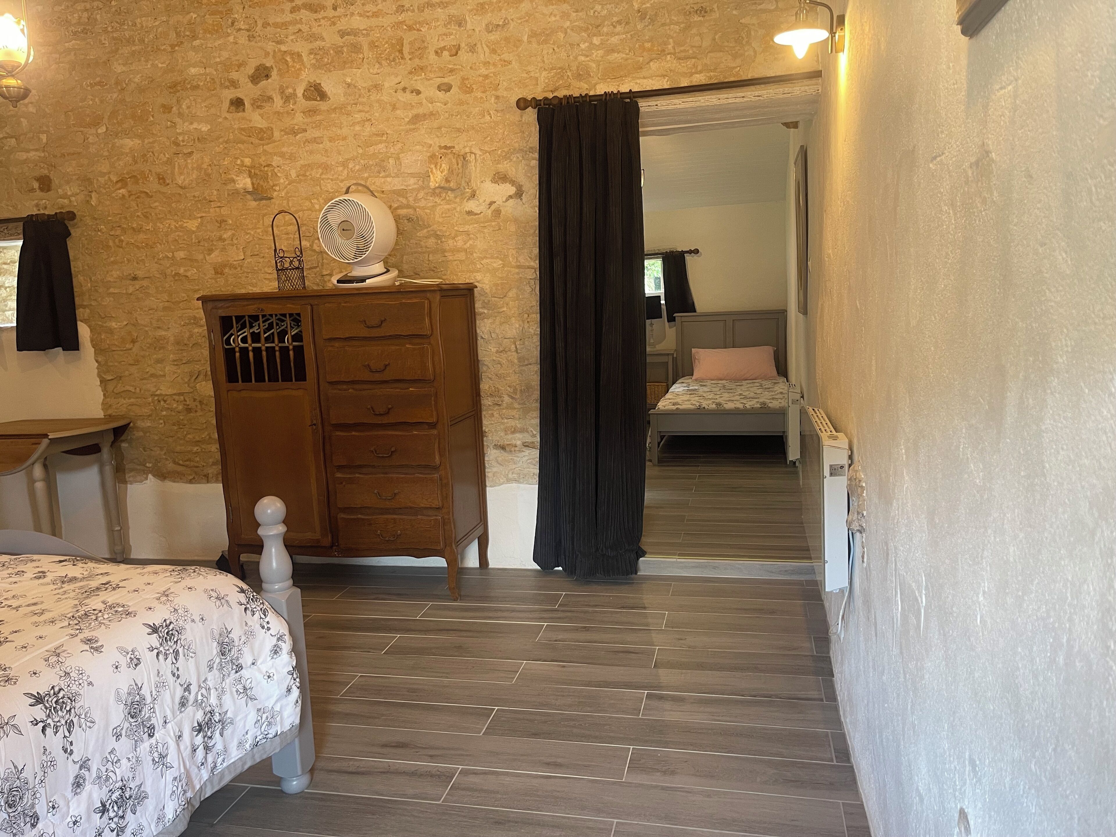 A bedroom with a wooden dresser, a bed with a floral bedspread, a nightstand, and a stone wall.
