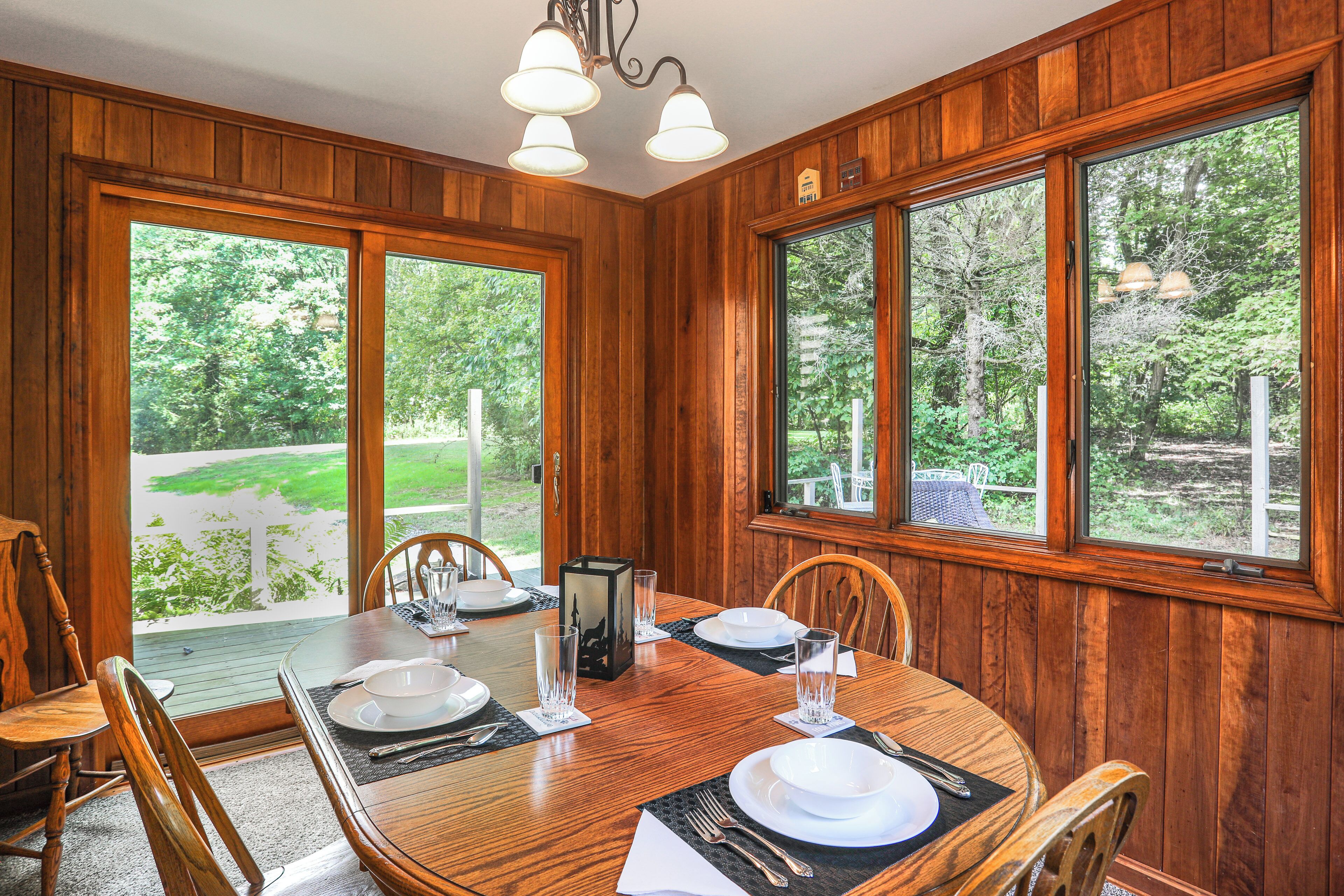 A dining area with a wooden table set for four, large windows, and a view of greenery outside.