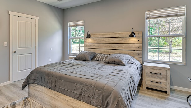 Un dormitorio con cabecera de madera, una cama grande con ropa de cama gris, dos mesitas de noche y vistas a árboles desde las ventanas.