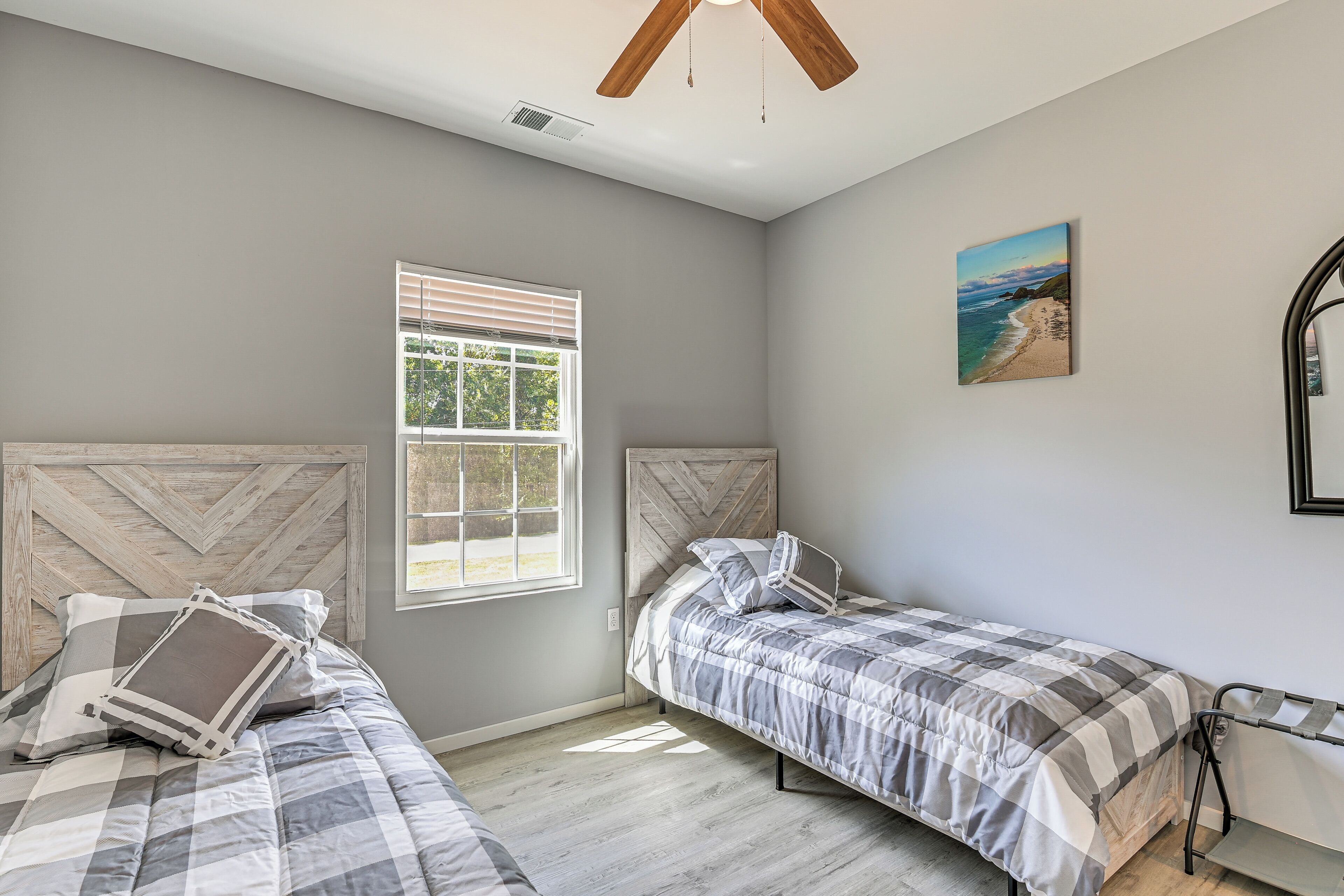 A bedroom with two beds, a window with blinds, a ceiling fan, and a beach painting on the wall.