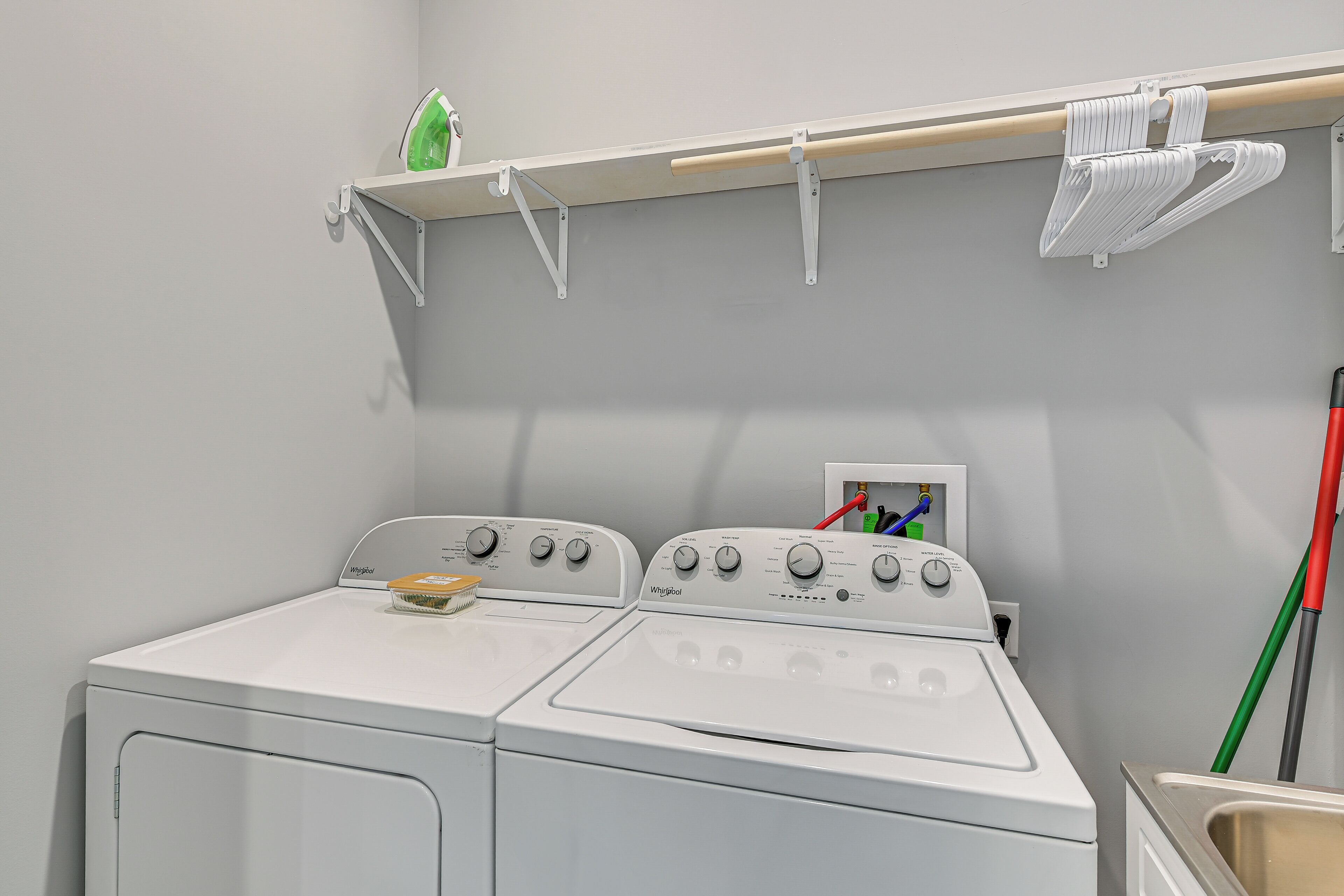 Une buanderie équipée d’un lave-linge et d’un sèche-linge à chargement frontal, d’une étagère sur laquelle se trouve une bouteille verte et d’un balai accroché au mur.