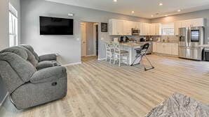 Una sala moderna con un sofá gris, un comedor con sillas blancas, una cocina con gabinetes blancos y electrodomésticos de acero inoxidable, y un televisor de pantalla plana montado en la pared.