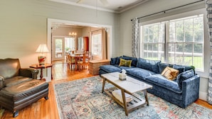 A spacious living room with a blue sofa, a leather armchair, a coffee table, and a patterned rug. There is a dining area visible through an open doorway. - 30 Acres & Pond: North Georgia Mtn Farmhouse (Demorest)