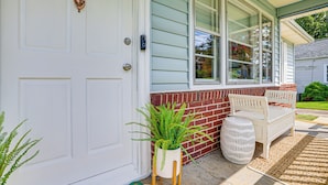 A white door with a brass knocker, a white bench, a potted plant, and a wicker basket outside a house with a brick foundation and a window with blinds. - Families Welcome, Fenced Yard: Inviting Greer Home (Greer)