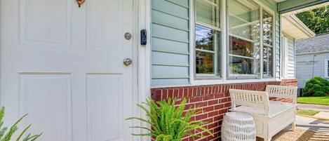 A white door with a brass knocker, a white bench, a potted plant, and a wicker basket outside a house with a brick foundation and a window with blinds.
