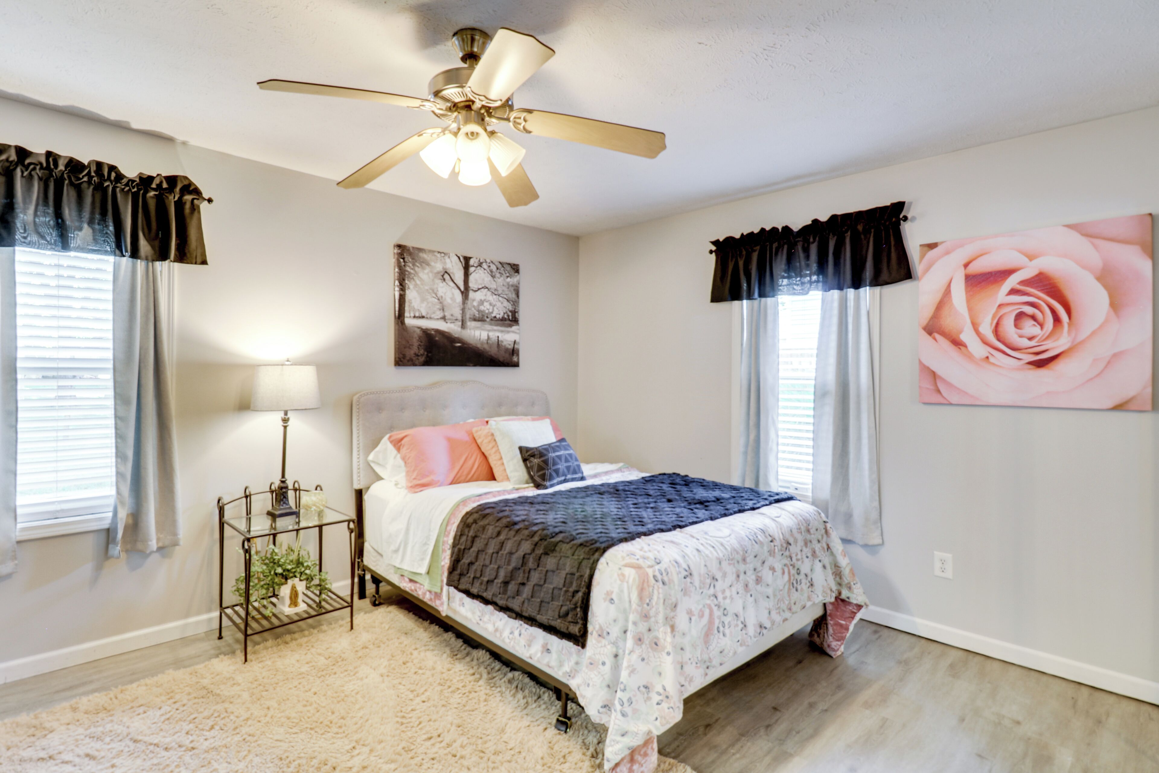 A bedroom with a bed, a ceiling fan, a nightstand, a lamp, and a large floral painting on the wall.