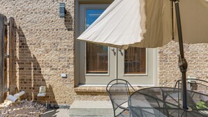 Un bâtiment en briques avec une porte et des fenêtres, une table en métal avec des chaises et un parasol blanc.
