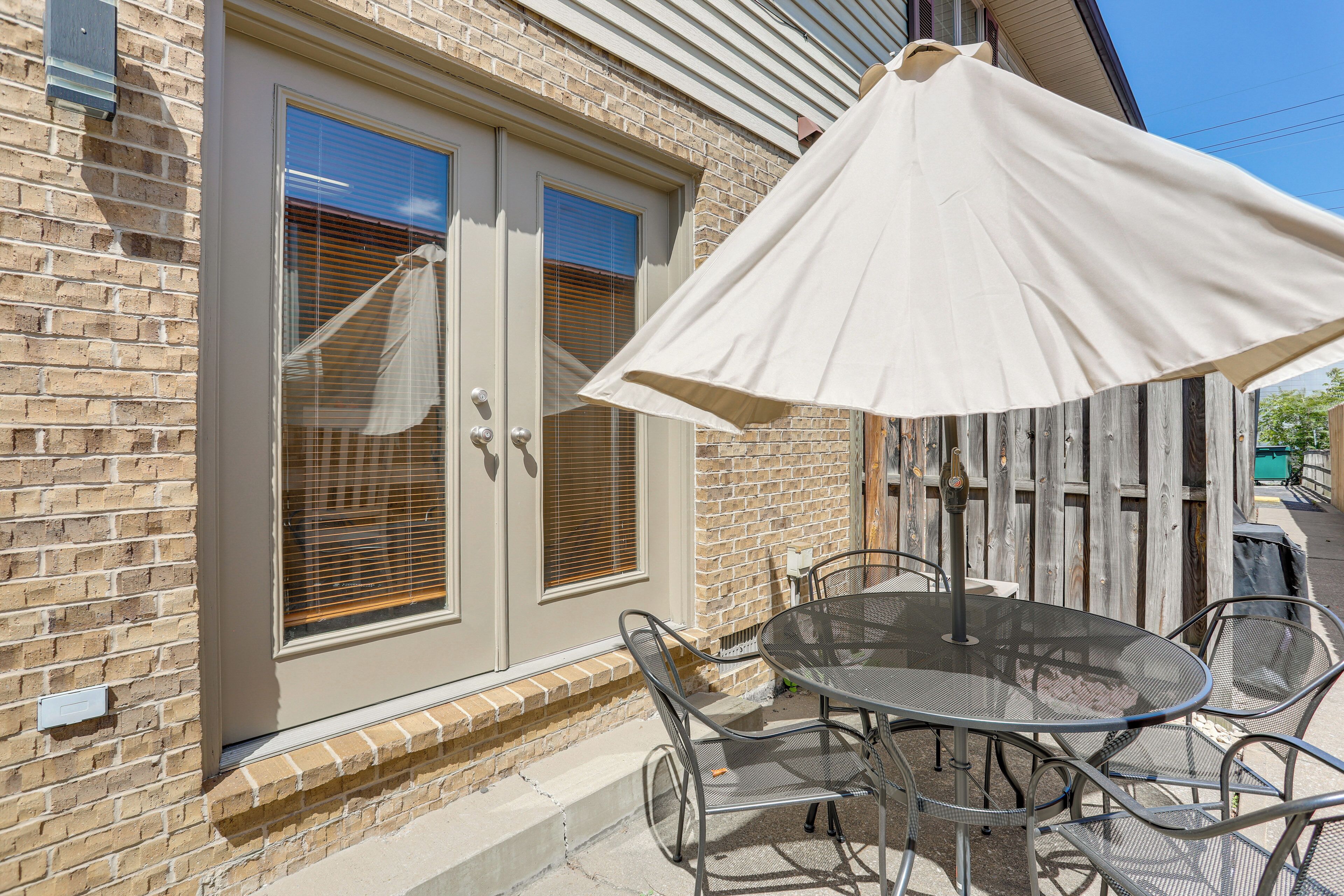 Eine Terrasse mit einem runden Tisch und Stühlen, einem weißen Sonnenschirm sowie einem Haus aus Ziegelsteinen mit Fenster und Tür.