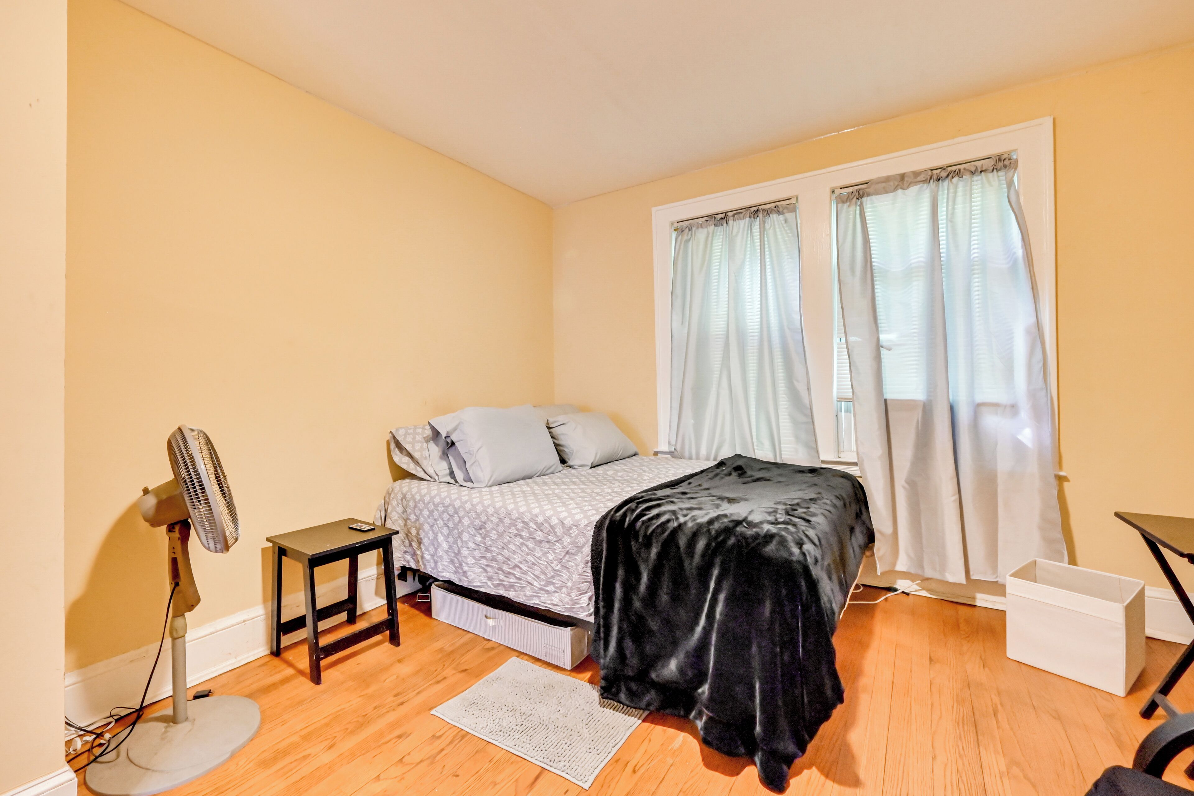 Une chambre à coucher avec un lit, un ventilateur sur pied, une petite table, un bureau et une fenêtre avec des rideaux.