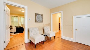 A hallway with two upholstered chairs, a wooden floor, and a door leading to a bedroom with a bed and curtains.