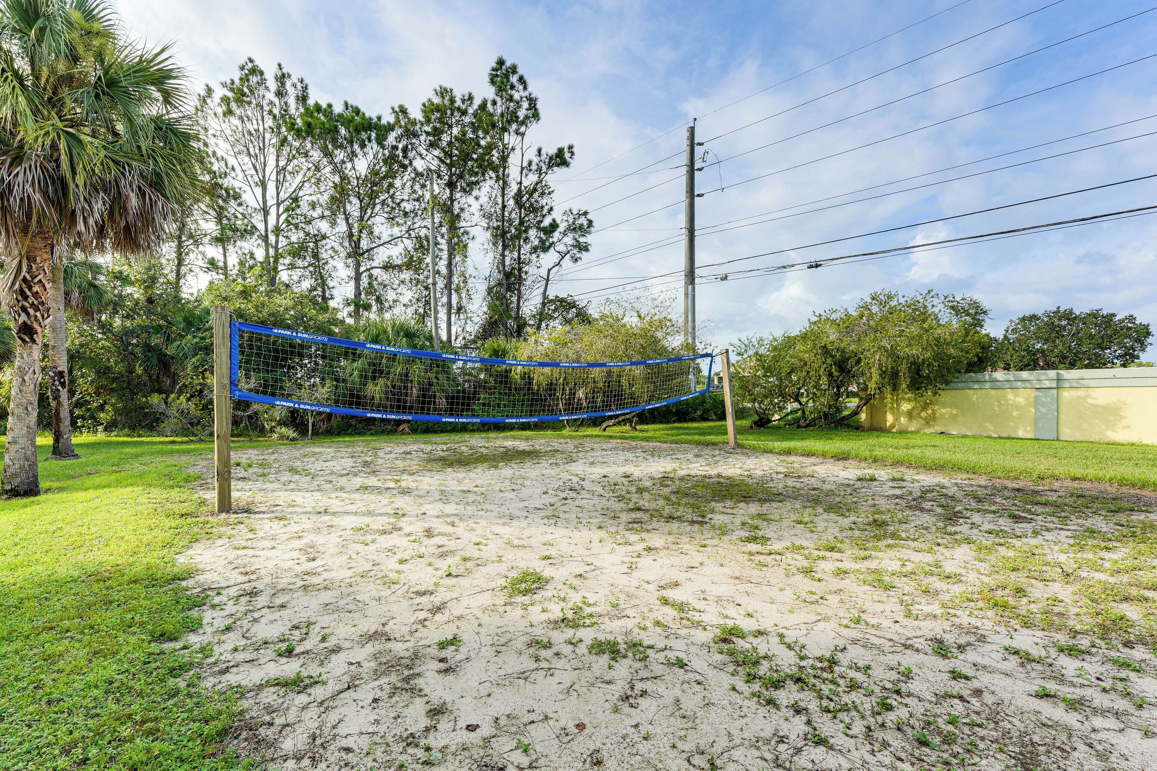 Una red de voleibol en una zona arenosa con árboles y una valla al fondo.