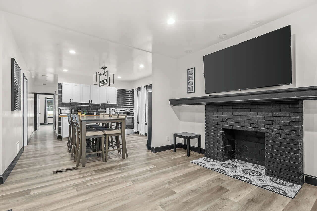 A modern kitchen with white cabinets, a dining area with a wooden table and chairs, and a black brick fireplace.