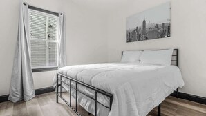 A bedroom with a bed, a window with curtains, and a cityscape painting on the wall.