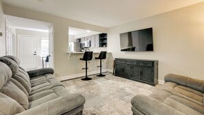 Un salon comprenant un canapé d’angle, deux tabourets de bar, un téléviseur à écran plat fixé au mur et un meuble en bois foncé.