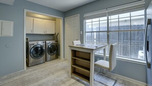 A laundry room with two front-loading washing machines, a table with a chair, and a window with blinds.