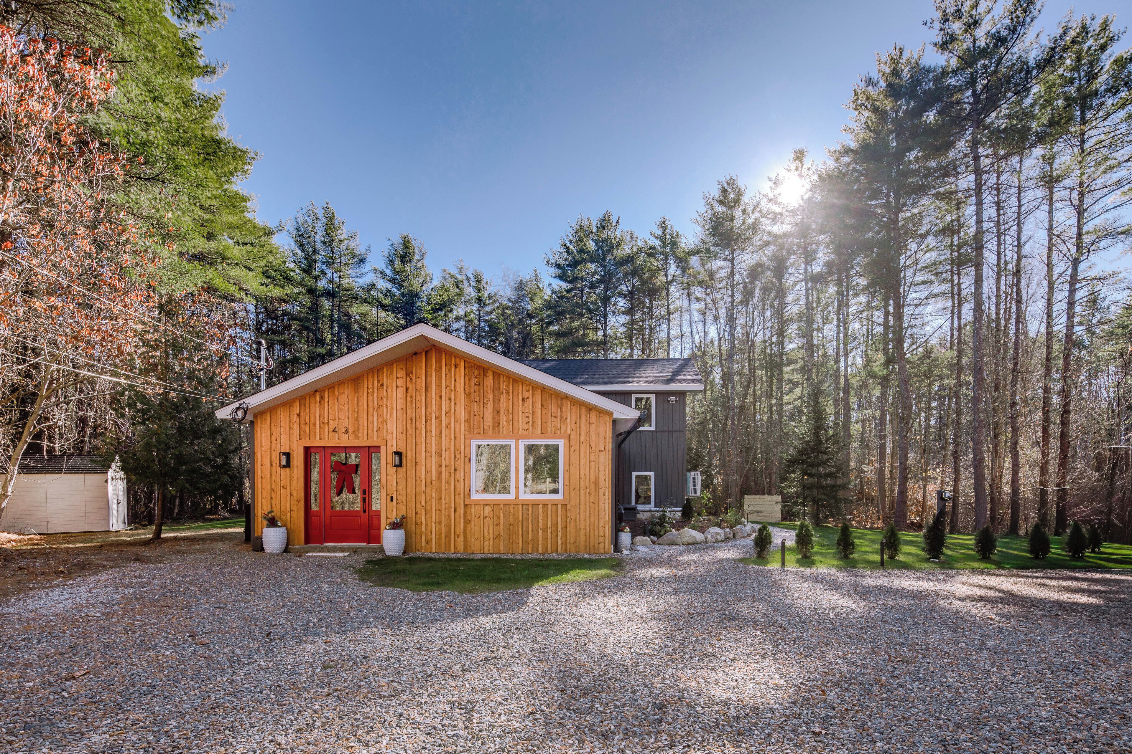A wooden cabin with a red door, surrounded by a gravel driveway and a forested area.