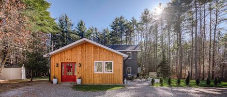 A wooden cabin with a red door, surrounded by a gravel driveway and a forested area.