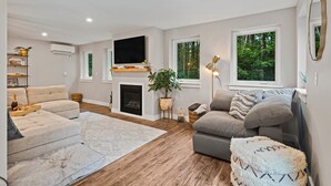 A modern living room with a fireplace, a flat-screen TV, and large windows offering a view of greenery. - Modern Gem on 15 Acres, 5 Mi to Old Orchard Beach! (Saco)
