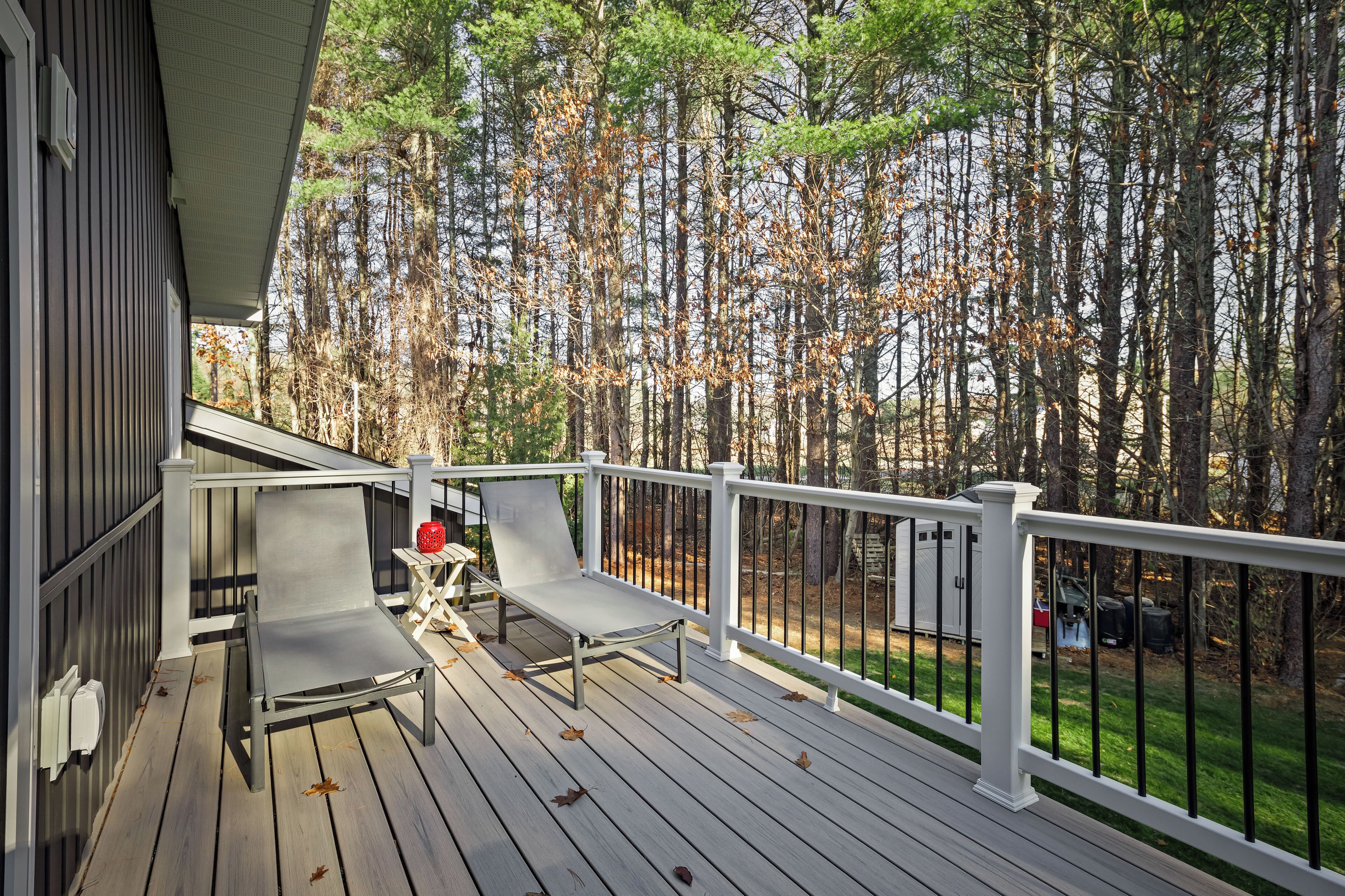 A wooden deck with two lounge chairs and a small table, surrounded by a white railing and a view of a forest.