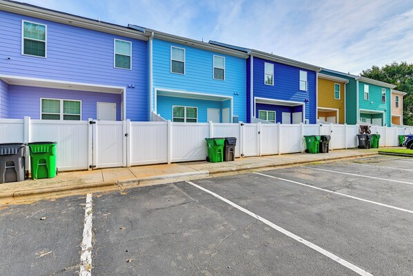 A row of colorful townhouses with white fences, parking spaces, and green trash bins.
