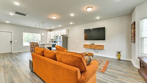 A modern living room with a sofa, a flat-screen TV mounted on the wall, and a kitchen in the background.