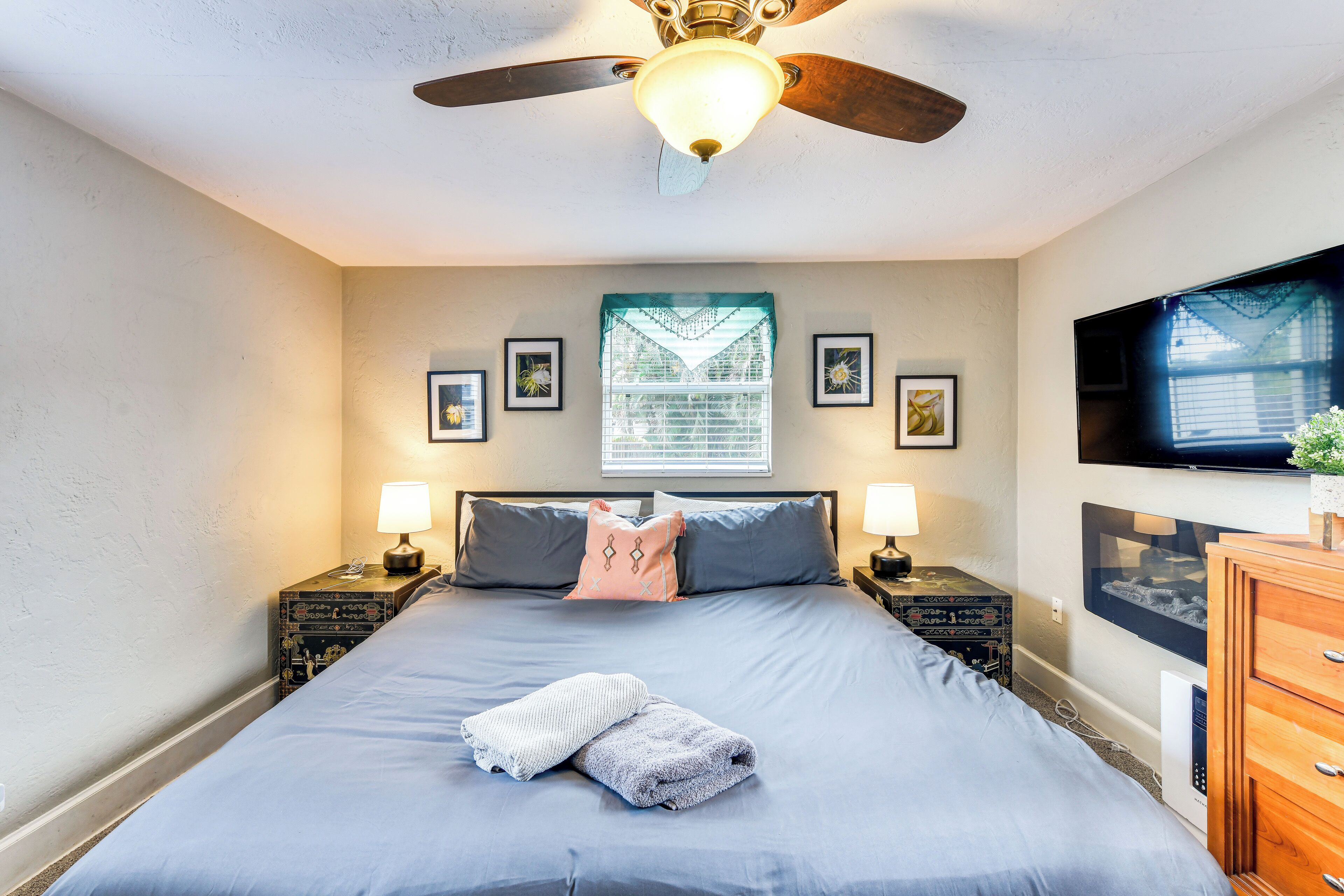 A neatly made bed with a blue bedspread, two pillows, and a folded towel. There are two bedside tables with lamps, a ceiling fan, and a flat-screen TV mounted on the wall.