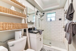 Un baño con inodoro, lavabo y ducha. Hay cestas de mimbre en estantes, un perchero y una ventana con vistas.