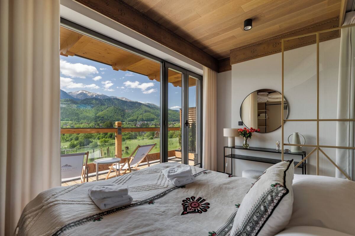 A bedroom with a large bed, a wooden ceiling, a balcony with a view of mountains, and a round mirror on the wall.