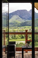 Un balcón con sillas de mimbre y una mesa, con vistas a un paisaje montañoso con vegetación y un pueblo abajo.