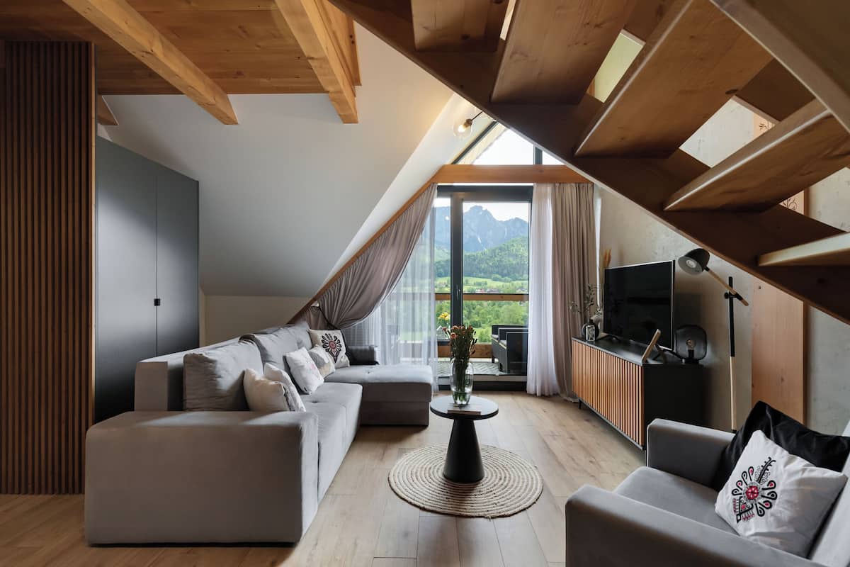 A modern loft bedroom with a sloped ceiling, a large window offering a mountain view, a built-in TV, and a cozy seating area with a sofa and armchair.