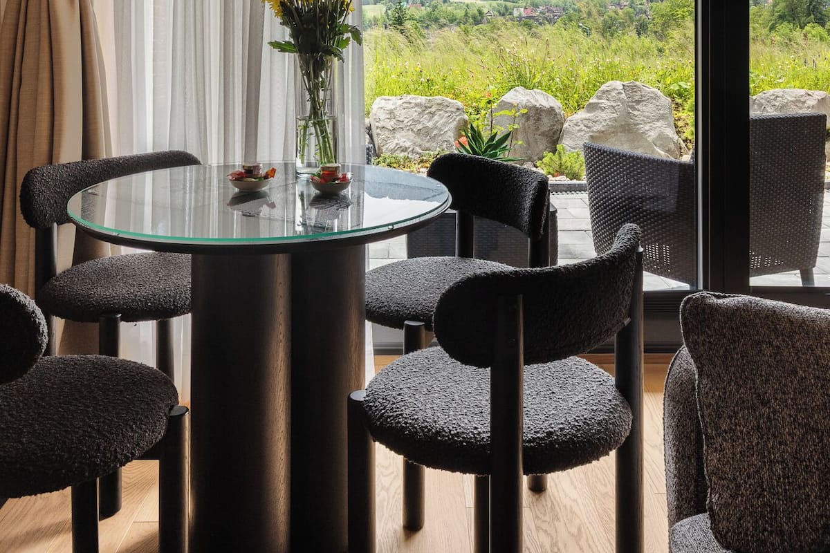 A dining area with a glass table, black chairs, a vase with yellow flowers, and a view of mountains through large windows.