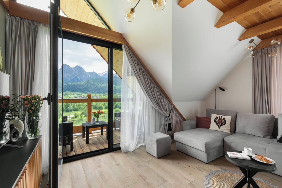 A modern loft bedroom with a large sliding glass door leading to a balcony, a grey sofa, and a small table with a book and a croissant.