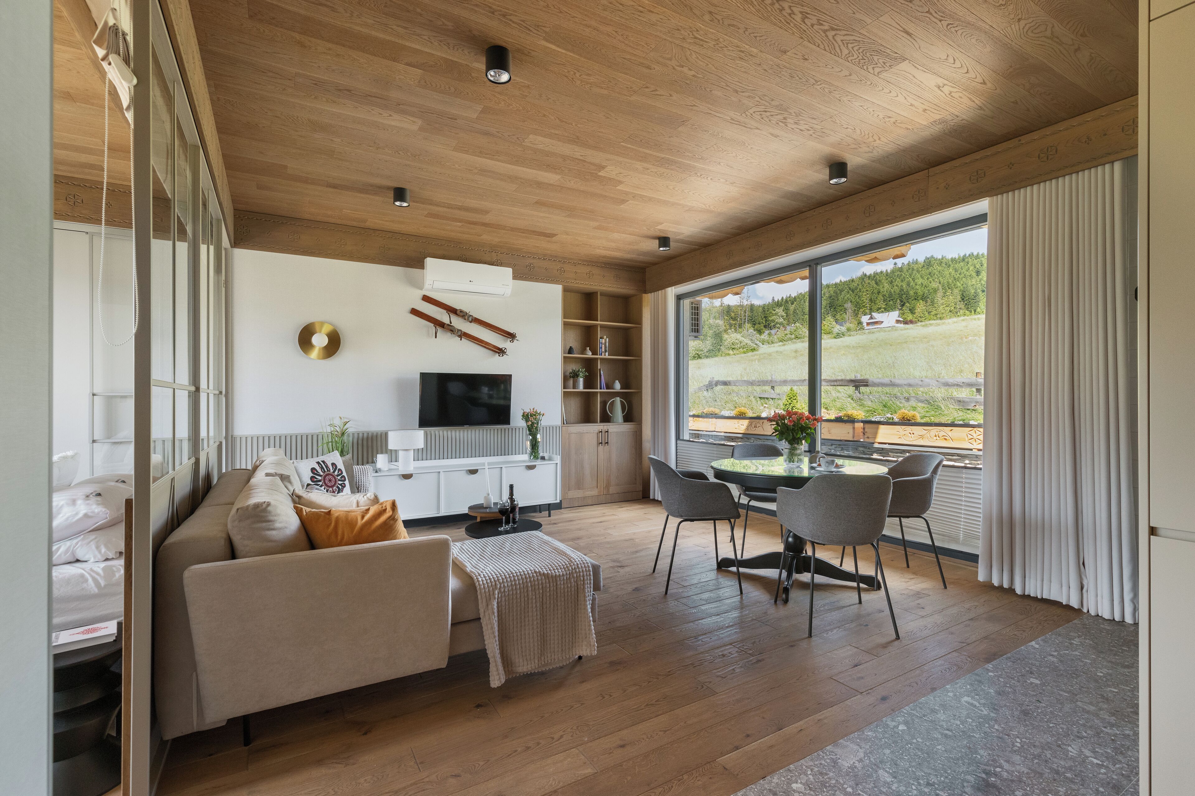 A modern living room with a wooden ceiling, a sofa, a TV, a dining area with chairs, and a large window with a view of greenery.