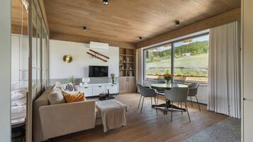 A modern living room with a wooden ceiling, a sofa, a TV, a dining area with chairs, and a large window with a view of greenery.