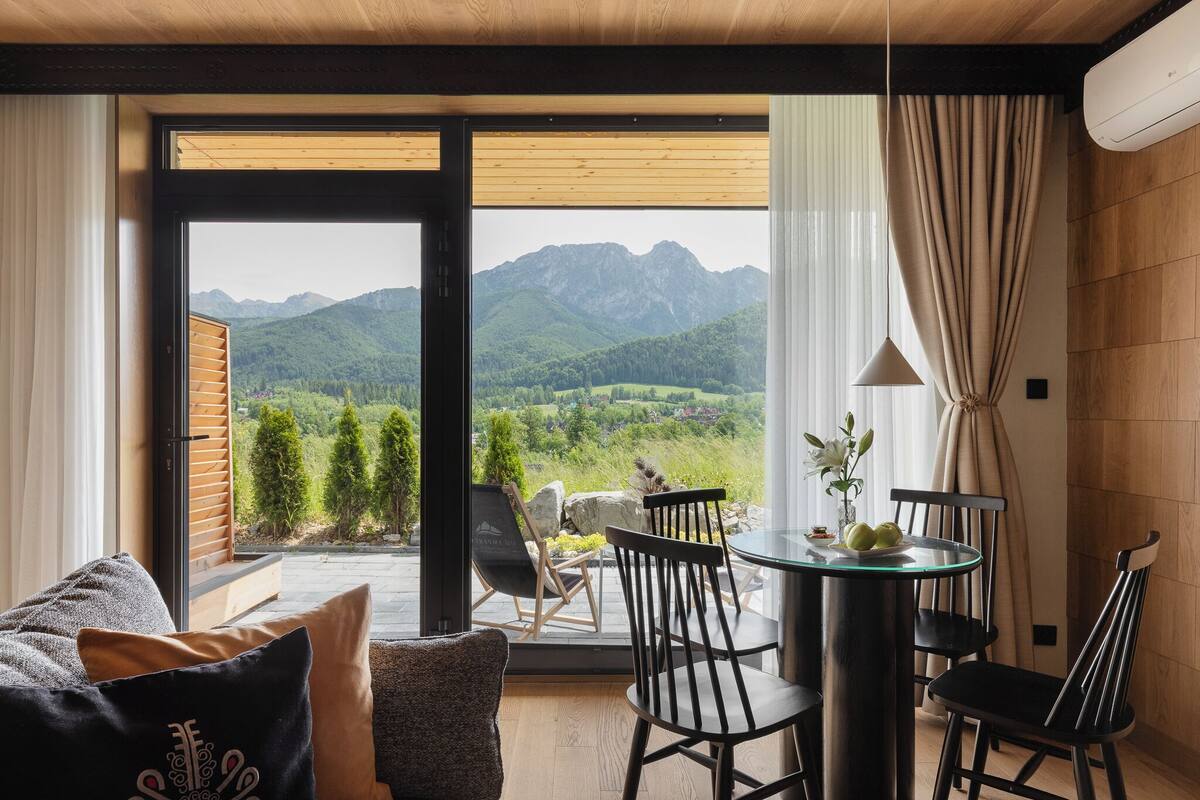 A room with a view of mountains, a dining table with chairs, and a sofa with cushions.