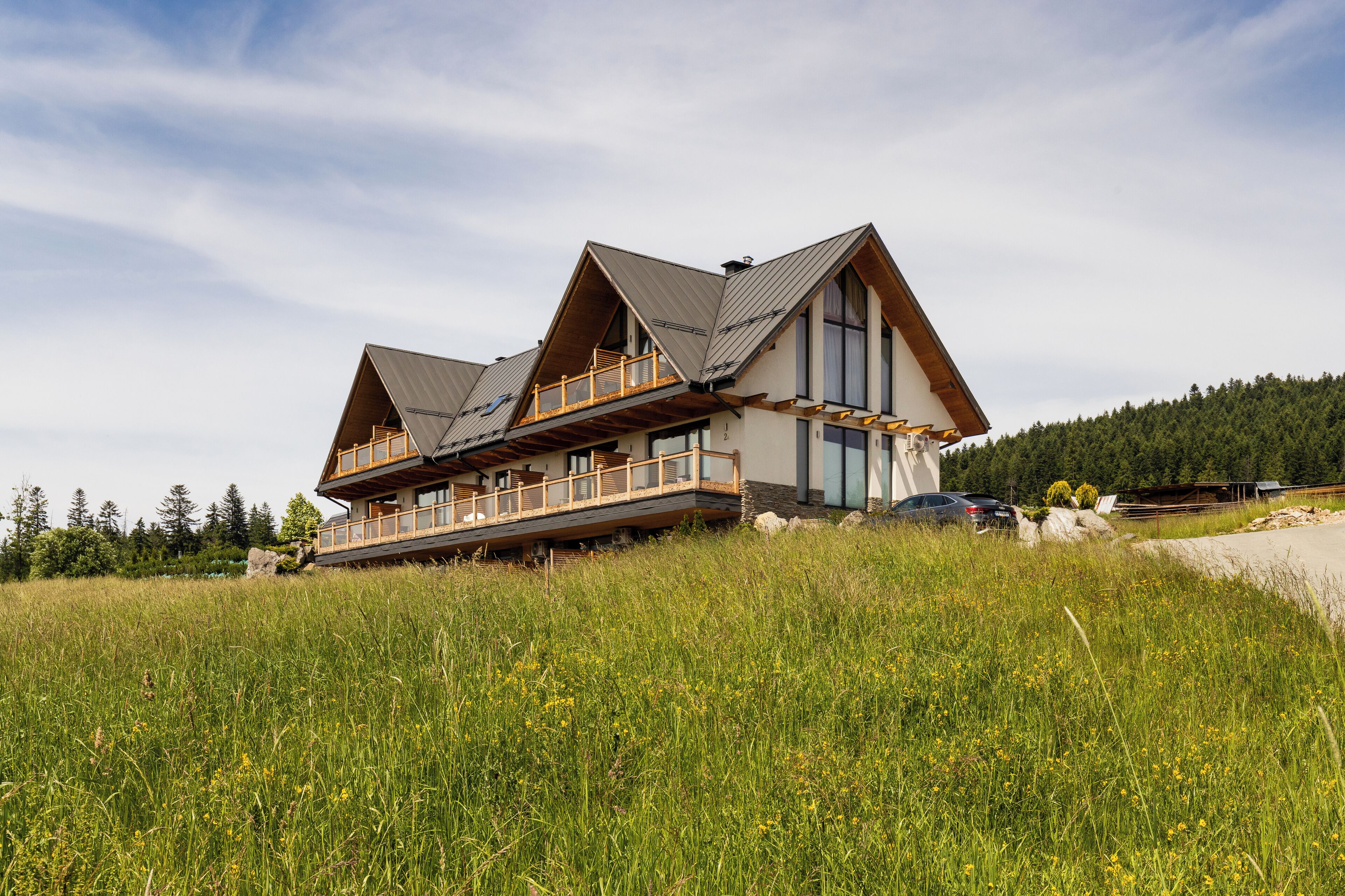 A modern house with a large balcony, situated on a grassy hill with a forest in the background.