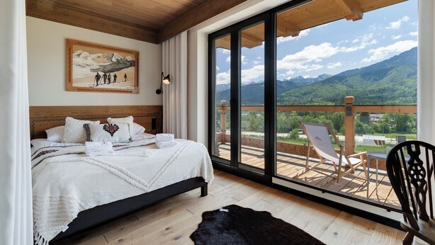 A bedroom with a large bed, a painting on the wall, and a balcony with chairs and a view of mountains. - IJG Imperial Apartamenty Szymaszkowa (Zakopane)