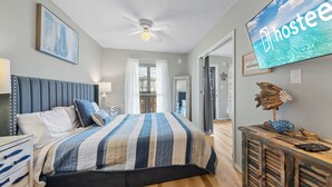 A bedroom with a bed, a ceiling fan, a TV displaying 'hostee', a wooden cabinet with decorative items, and a view of the outdoors through a window. - The Cove Condos (Gulf Shores)