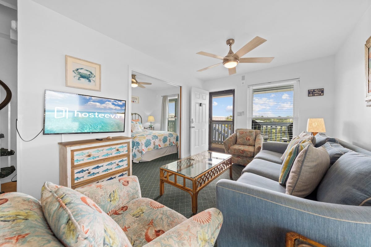 A living room with a television, a ceiling fan, a sofa, a chair, and a bed with a view.