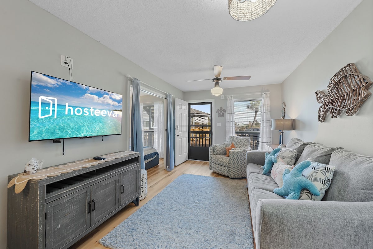 A living room with a television displaying 'hosteeva', a grey sofa with starfish pillows, a wooden cabinet, and a blue rug.