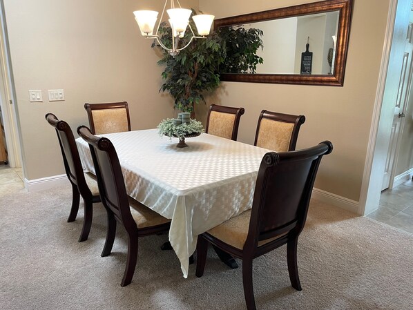 A dining room with a rectangular table, six chairs, a chandelier, a potted plant, and a mirror on the wall.