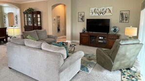 Ein Wohnzimmer mit grauem Sofa, braunem Sessel, Fernseher auf Holzständer und Deckenventilator.