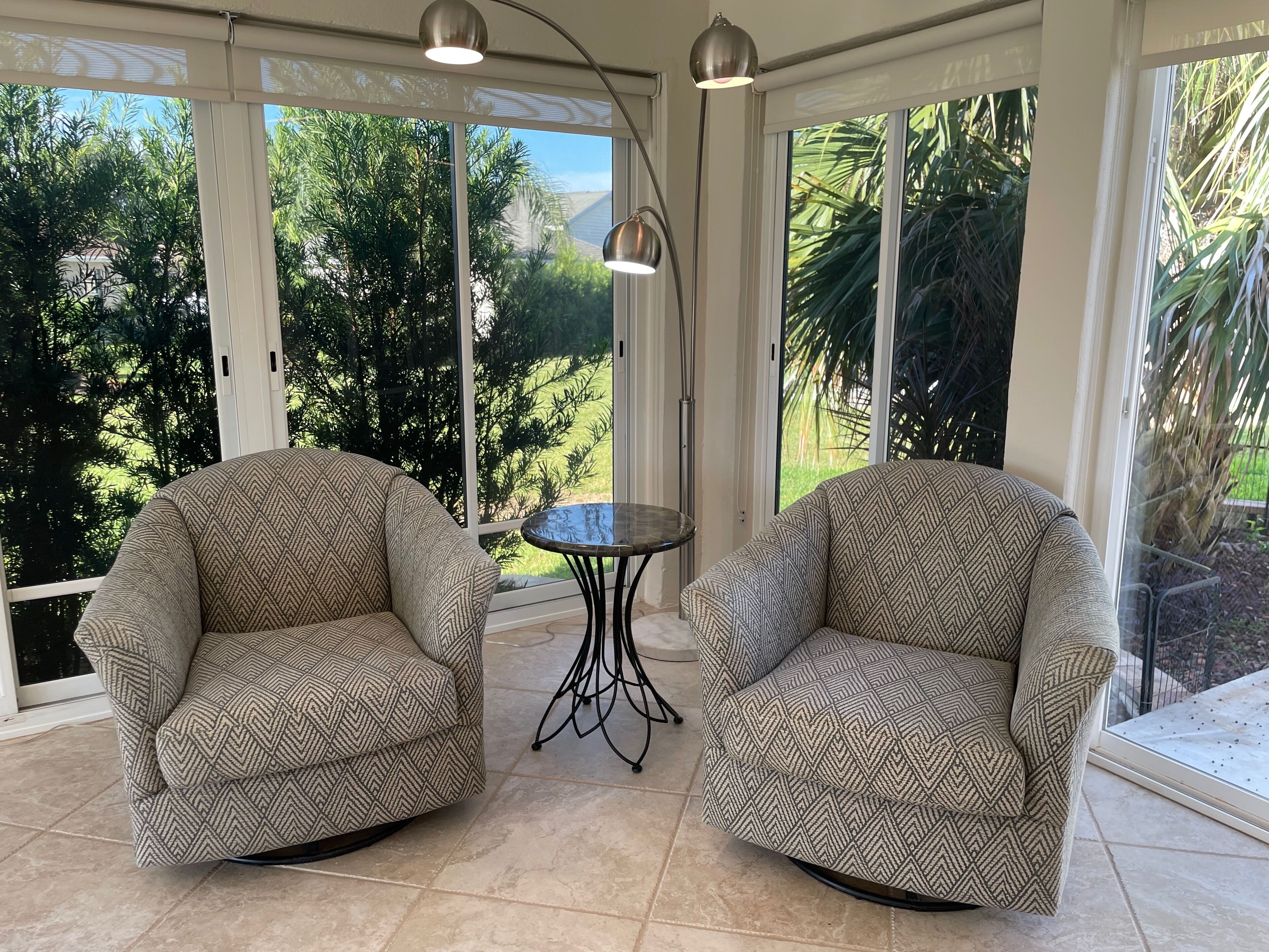 A room with two patterned armchairs, a small round table, and large windows offering a view of greenery.