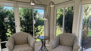 A room with two patterned armchairs, a small round table, and large windows offering a view of greenery.