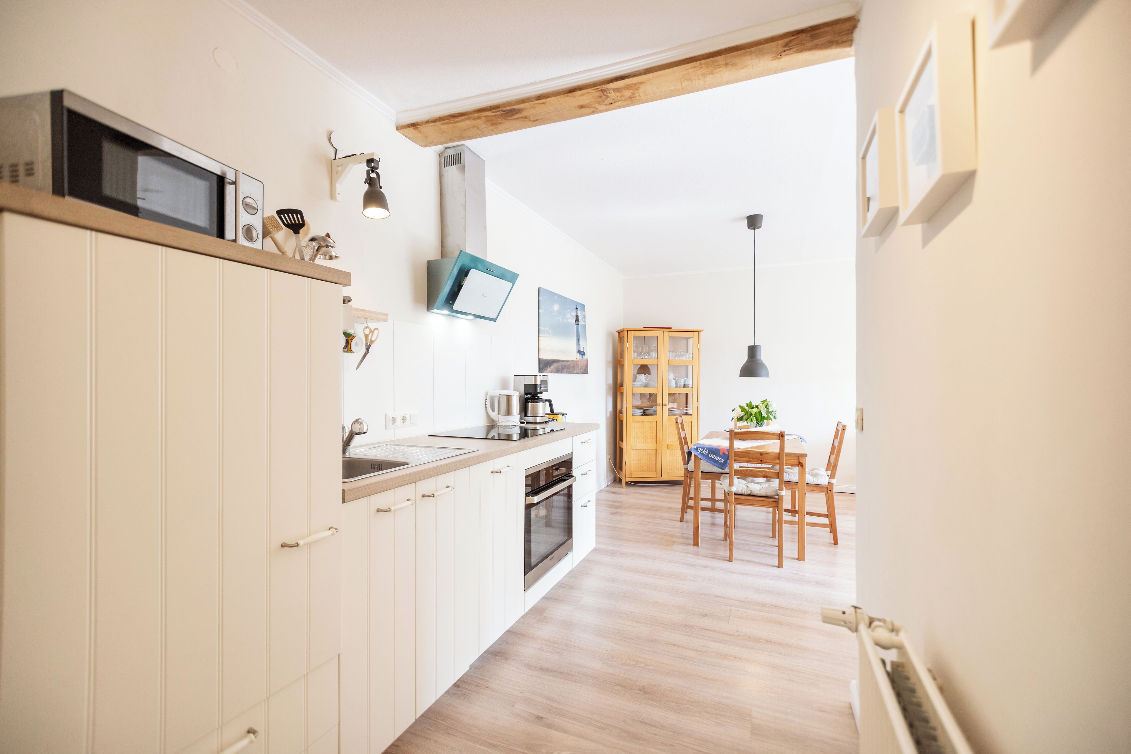 Eine moderne Küche mit weißen Schränken, einer Mikrowelle und einer Kaffeemaschine. Ein Holztisch mit Stühlen lädt zum Essen ein, und durch eine offene Tür ist der Wohnbereich sichtbar.