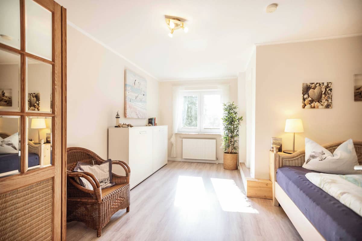 Habitación luminosa y amplia con una cama grande, una silla de mimbre, una mesita con una lámpara, una planta en maceta y vistas al exterior a través de una ventana.
