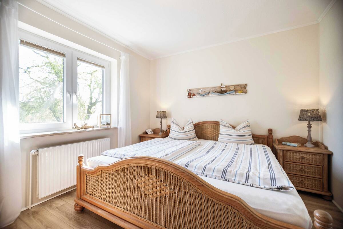 Un dormitorio con cama de madera, ropa de cama blanca, ventana con persianas, mesita de noche con lámpara y una obra de arte colgada en la pared.