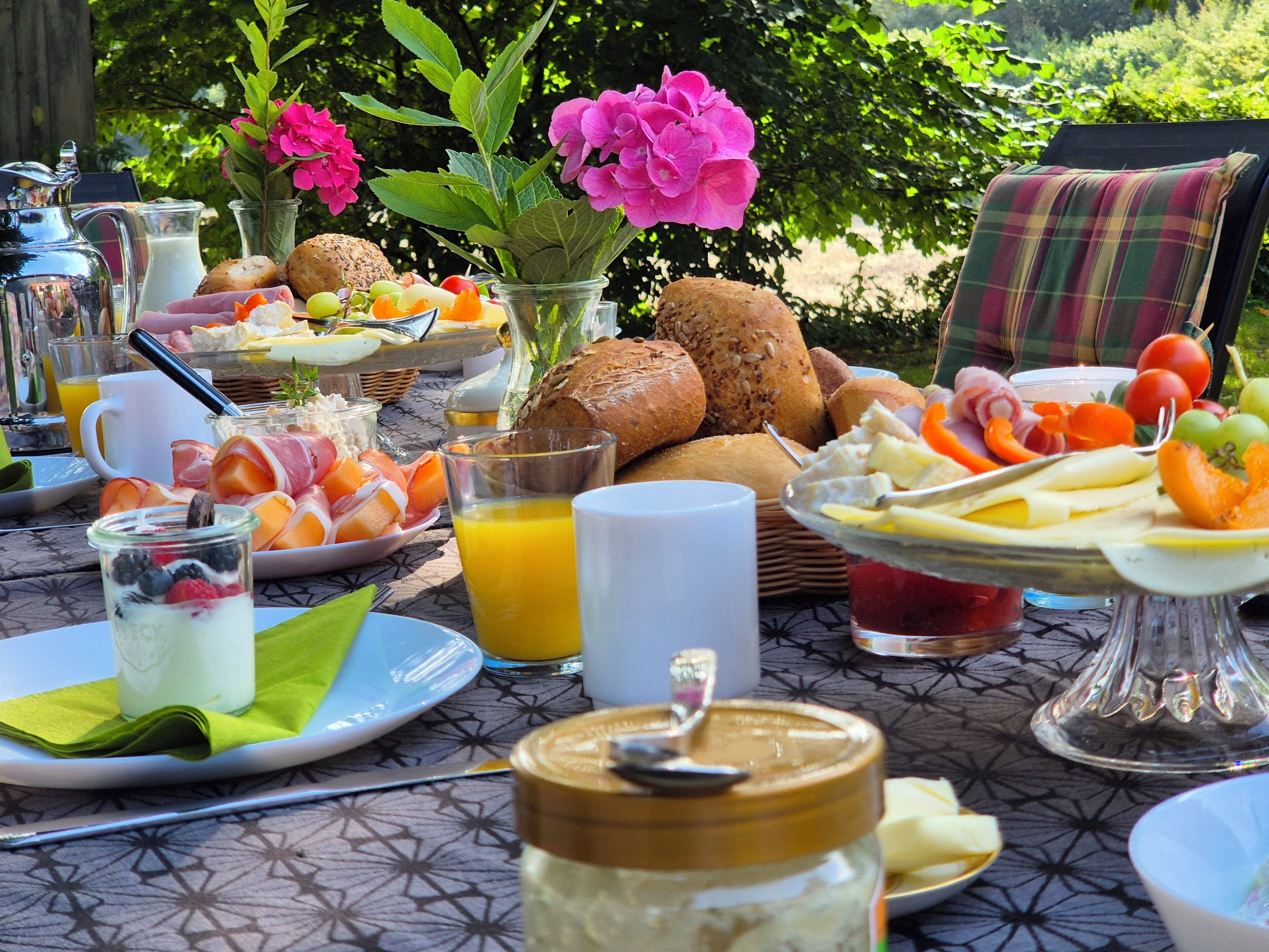 A breakfast table with various food items, a glass of orange juice, and a vase with flowers.